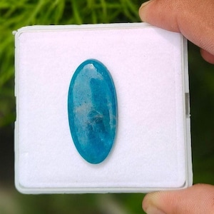 May include: An oval-shaped, translucent blue gemstone with a smooth, polished surface. The stone displays variations in colour intensity and internal clarity, resting centred on a square white display pad within a clear plastic storage case.