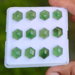 May include: A collection of twelve faceted, hexagonal green gemstones arranged in a grid pattern inside a white display box. Each stone features a geometric cut with multiple triangular facets that reflect light across their translucent surfaces.