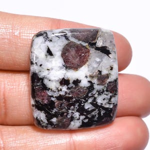 Puede incluir: Una piedra preciosa rectangular con una superficie pulida. La piedra presenta una mezcla de colores blanco, negro y burdeos. El burdeos aparece en pequeñas inclusiones redondas. El patrón general es una mezcla de texturas moteadas y veteadas.