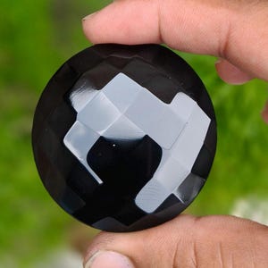 Könnte beinhalten: Ein runder, schwarzer Edelstein mit facettierter Oberfläche. Der Stein hat eine polierte Oberfläche, die das Licht über seine zahlreichen geometrischen Ebenen reflektiert und ein kontrastreiches Erscheinungsbild mit tiefschwarzen Tönen und hellen weißen Lichtreflexen erzeugt.