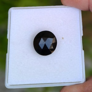 Könnte beinhalten: Ein runder, facettierter schwarzer Edelstein in der Mitte einer quadratischen weißen Schmuckschatulle. Der Stein hat eine polierte Oberfläche mit geometrischen Schliffen, die das Licht reflektieren und deutliche dunkle Facetten erzeugen.