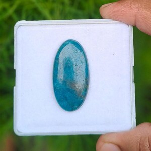 Könnte beinhalten: Ein oval geschliffener, blauer Edelstein mit sichtbaren Einschlüssen und einer glatten, reflektierenden Oberfläche. Der Stein ist mittig auf einer quadratischen, weißen Präsentationskarte platziert und zeigt eine lebendige blaugrüne Farbe.