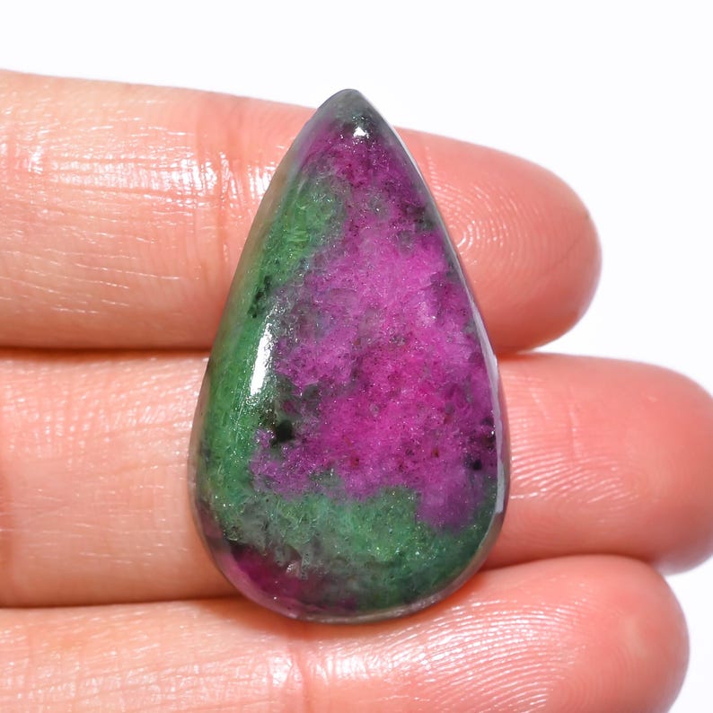 Puede incluir: Una gema en forma de l&aacute;grima con una superficie pulida. La piedra presenta una vibrante mezcla de colores, incluyendo magenta intenso, verde bosque y toques de negro. La gema se sostiene en una mano.