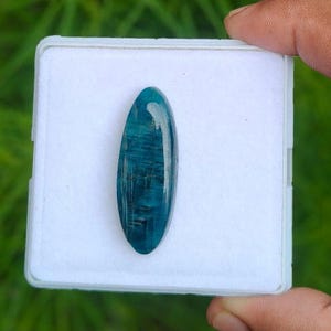 Könnte beinhalten: Ein polierter, oval geformter blauer Edelstein mit einer glatten Oberfläche im Cabochon-Stil. Der Stein zeigt tiefe blaugrüne und marineblaue Töne mit sichtbaren inneren faserigen Einschlüssen und einem glänzenden Finish auf einem weißen Pad.
