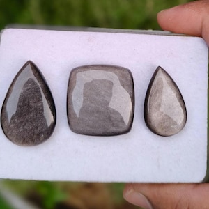 Könnte beinhalten: Drei polierte Silberglanz-Obsidian-Edelsteine auf einer weißen rechteckigen Karte. Das Set enthält zwei tropfenförmige Steine und einen quadratischen Stein mit abgerundeten Ecken, alle mit metallisch grauem Glanz und glatter Oberfläche.