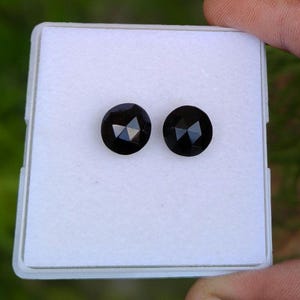 May include: Two round, faceted black gemstones displayed on a white square display pad. The stones feature a rose-cut style with multiple triangular facets that reflect light, creating a dark, polished appearance against the plain white background.