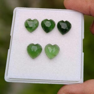 May include: Five heart-shaped green gemstones with faceted surfaces arranged in a white display box. The stones vary in shade from light to dark green, each featuring a polished finish and a distinct heart silhouette with a pointed base and top notch.