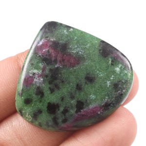 Może przedstawiać: Wypolerowany, zaokrąglony kamień szlachetny o zielonej barwie podstawowej, nakrapiany czernią i plamami głębokiej czerwieni. Kamień trzymany jest w dłoni, ukazując jego gładką, naturalną fakturę i unikalny wzór kolorystyczny. Naturalny kamień szlachetny.