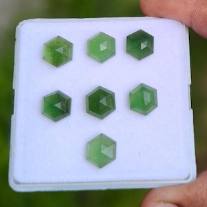 May include: Seven loose, hexagonal-cut green gemstones arranged on a white square display pad. Each stone features a faceted surface with a translucent appearance, displaying varying shades of deep and light green throughout the collection.