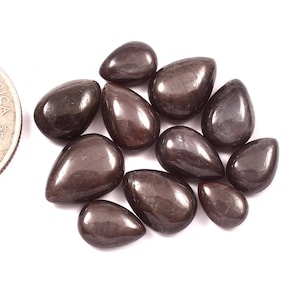 May include: A collection of polished, dark brown gemstones with a teardrop shape. The stones have a glossy, reflective surface and are arranged on a white background. The gemstones vary in size and shape.