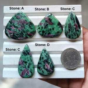 Könnte beinhalten: Fünf polierte Zoisit-Steine mit Rubineinschlüssen, beschriftet als Stone A, B, C und D, auf einem weißen Display angeordnet. Die Steine zeigen grüne, schwarze und magentafarbene Muster. Eine silberfarbene Münze liegt daneben als Größenvergleich.