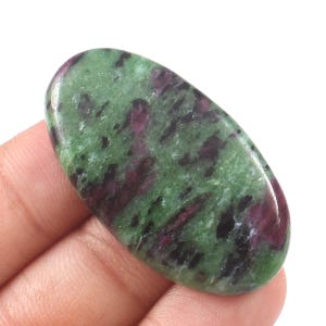 Könnte beinhalten: Ein ovaler Edelstein mit polierter Oberfläche. Der Stein weist ein gesprenkeltes Muster aus tiefen Grün-, Dunkelrot- und Schwarztönen auf. Der Edelstein wird in einer Hand vor weißem Hintergrund gehalten.