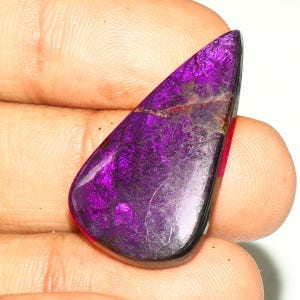 Puede incluir: Una piedra preciosa en forma de pera con un tono púrpura intenso y vibrante y una superficie pulida y reflectante. La piedra muestra una red compleja de vetas oscuras e intrincadas e inclusiones metálicas que crean una textura.