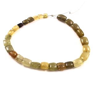 Może przedstawiać: Naszyjnik z kwadratowych kamieni szlachetnych w odcieniach żółci, brązu i zieleni. Koraliki są nawleczone na cienki drut, tworząc zakrzywiony kształt na białym tle. Kamienie szlachetne mają wypolerowaną, gładką powierzchnię.