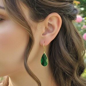 Könnte beinhalten: Goldfarbene Ohrhänger mit einem facettierten, smaragdgrünen, tropfenförmigen Edelstein. Die Ohrringe hängen an einem zarten goldenen Haken und zeigen ein klassisches und elegantes Design. Die Ohrringe werden am Ohr einer Person gezeigt.