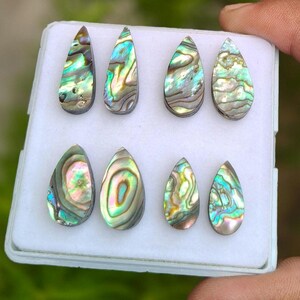 May include: Eight teardrop-shaped abalone shell cabochons arranged in two rows on a white display tray. Each piece features a unique iridescent surface with swirling patterns of green, blue, pink, and silver tones, showcasing natural shell textures.