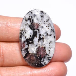 Może przedstawiać: Owalny kamień szlachetny z nakrapianą białą podstawą, poprzecinaną czarnymi i ciemnoczerwonymi wtrąceniami. Wypolerowana powierzchnia kamienia odbija światło, podkreślając naturalne wzory i tekstury. Z bliska widać unikalny skład mineralny.