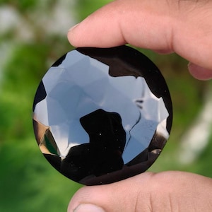 Könnte beinhalten: Ein runder, facettierter schwarzer Edelstein mit einer polierten, reflektierenden Oberfläche. Der Stein weist mehrere geometrische Schliffe auf, die scharfe Kanten und lichtreflektierende Flächen auf seinem dunklen, undurchsichtigen Körper erzeugen.