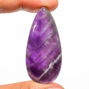 Könnte beinhalten: Ein polierter, tropfenförmiger Amethyst-Edelstein. Der Stein hat einen tiefvioletten Farbton mit helleren violetten und weißen Chevron-Mustern. Der Edelstein wird zwischen zwei Fingern vor einem weißen Hintergrund gehalten.