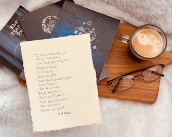 May include: A still-life arrangement featuring a handwritten poem on textured paper, alongside notebooks with celestial designs, a cup of coffee, and eyeglasses. The poem's text is visible. The items are arranged on a wooden board.