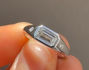 May include: A silver ring featuring a rectangular, clear gemstone at the center, flanked by two smaller, round gemstones. The ring has a modern, minimalist design, with a polished finish. The ring is held by a person's fingers.
