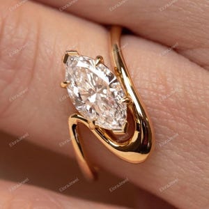 May include: A marquise-cut diamond engagement ring with a unique, flowing gold band. The ring's design features a central, sparkling diamond held by delicate prongs, set in a polished gold setting. The band curves gracefully around the finger.