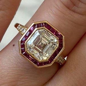 Op de afbeelding: Een gouden ring met een grote, vierkant geslepen diamant in het midden, omringd door een halo van kleine, rechthoekige robijnen. De ring heeft ook diamantaccenten op de band. Dit is een sieraad.