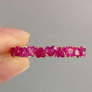May include: A gold band ring featuring a series of heart-shaped, vibrant pink gemstones. The gemstones are closely set, creating a continuous row of hearts. The ring is held by a person's fingers against a neutral gray background.