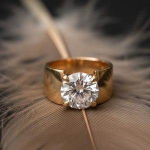 May include: A gold ring featuring a large, clear, round gemstone. The ring rests on a brown feather against a dark backdrop. The ring's band is wide and smooth, with the gemstone secured by four prongs.