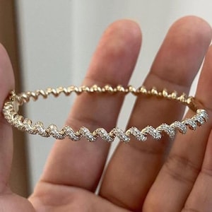 May include: A gold-toned bangle bracelet with a wavy design. The bracelet is encrusted with small, sparkling white gemstones. The bracelet is held in a person's hand, showcasing its intricate details and elegant style.