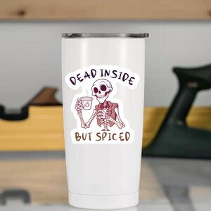 Może przedstawiać: Biały kubek podróżny z zabawną grafiką szkieletu trzymającego filiżankę kawy. Tekst na kubku brzmi "DEAD INSIDE BUT SPICED" w zabawnym kroju pisma. Szkielet ma na sobie brązową kamizelkę.