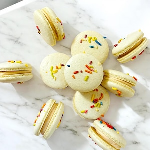 May include: A collection of nine macarons with pastel yellow shells and white frosting. The macarons are decorated with colorful sprinkles.