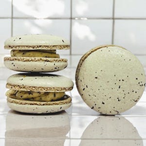 May include: A stack of three light green macarons with a light brown filling. The macarons have a speckled texture. A single macaron is shown to the right of the stack.