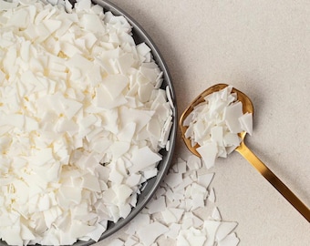 May include: White wax flakes in a gray bowl and a gold spoon. The wax flakes are scattered on a light gray surface.