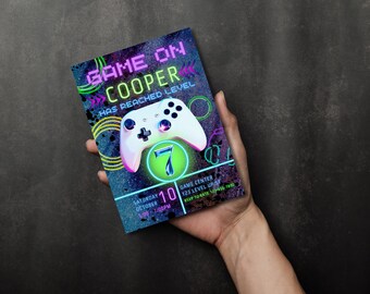 以下が含まれることがあります： ビデオゲームをテーマにした誕生日パーティーのネオンカラーの招待状。招待状には、緑色の光る数字7がついたビデオゲームコントローラーが描かれています。招待状のテキストは「Game On Cooper Has Reached Level 7」です。