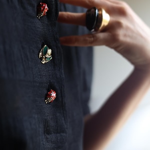 Può includere: Primo piano di un top in maglia nero con tre bottoni decorativi. I bottoni sono dorati con dettagli in smalto rosso e verde. È visibile anche un grande anello dorato con una pietra scura.