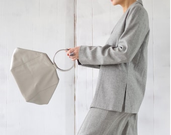 May include: A light gray skirt suit with a matching handbag. The suit includes a blazer and a long skirt. The handbag has a geometric shape and a silver handle. The person is wearing gray boots and a green beret.