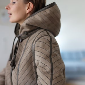 Può includere: Un cappotto con cappuccio beige con un motivo a righe diagonali e finiture marrone scuro. Il cappotto ha una chiusura con coulisse e un cappuccio. Le maniche sono lunghe e il cappotto sembra essere fatto di un materiale morbido.