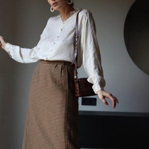 Pode incluir: Uma mulher vestindo uma blusa branca com uma saia marrom e branca de pata de galo e botas de couro marrom. Ela está carregando uma bolsa de ombro de couro marrom.