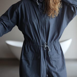 May include: A dark blue hooded jacket with a drawstring waist and pockets. The jacket has a relaxed fit and a unique, slightly ballooned shape at the bottom. The fabric has a subtle vertical stripe pattern.