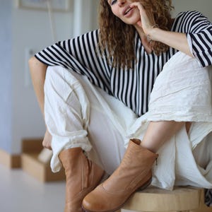 May include: A person wearing a black and white striped top and white pants, sitting on a wooden stool. The person is wearing light brown ankle boots with zippers.