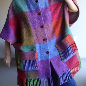Op de afbeelding: Een kleurrijke poncho-achtige jas met een geruit patroon, een knoopsluiting aan de voorkant en twee voorzakken met franjes. De jas heeft een levendige mix van kleuren, waaronder paars, rood, blauw, groen en oranje. De jas is gemaakt van een dikke, geweven stof.