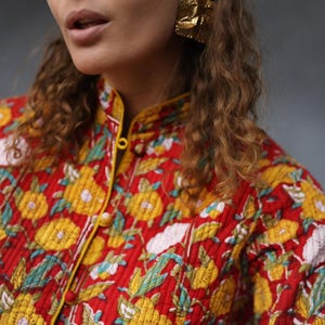 May include: A red quilted shirt with a yellow floral pattern and a mandarin collar. The shirt has a yellow trim and buttons down the front.