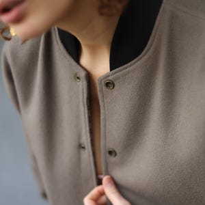 May include: A close-up of a taupe bomber jacket with a black collar and snap closures. The jacket has a soft, textured appearance. The collar is black, and the jacket is buttoned at the top.