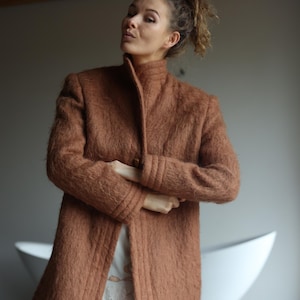 May include: A long, russet-coloured coat with a high collar and button closure. The coat has a textured, fuzzy appearance and features detailed stitching along the edges and sleeves. The coat is worn over a white dress.
