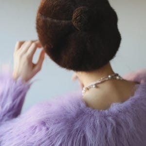 May include: A brown, fuzzy beret with a small pom-pom detail is worn with a lavender faux fur jacket. A delicate pearl necklace is visible. The image is a back view, focusing on the textures and colors of the clothing.