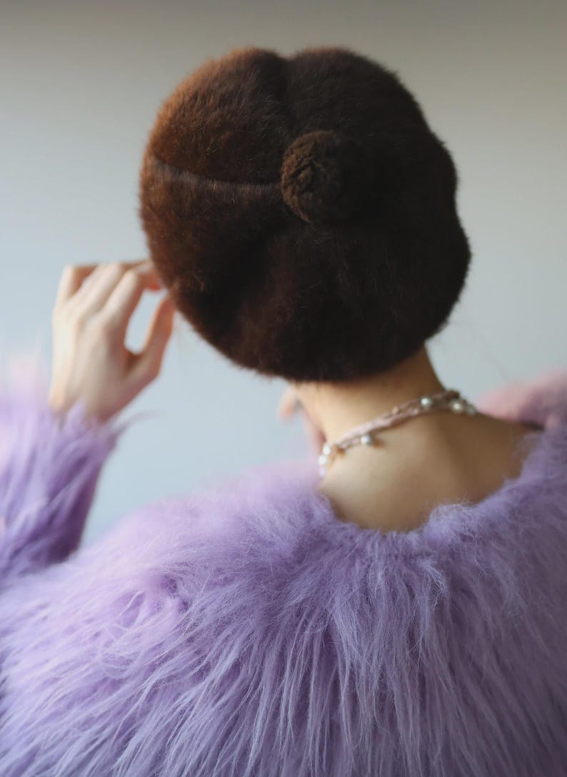 May include: A brown, fuzzy beret with a small pom-pom detail is worn with a lavender faux fur jacket. A delicate pearl necklace is visible. The image is a back view, focusing on the textures and colors of the clothing.