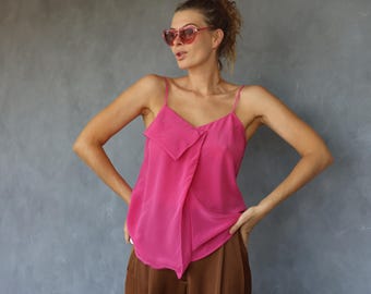 Pode incluir: Um top camisola rosa brilhante com alças finas e uma frente drapeada. O top é combinado com calças castanhas. A modelo usa óculos de sol rosa e tem o cabelo preso.