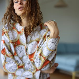 May include: A button-up shirt with a white background and a repeating pattern of gold-rimmed clocks. The shirt has orange accents and long sleeves. The model is wearing blue jeans.