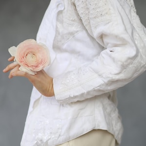 May include: White jacket with intricate embroidery and embellishments. The jacket features detailed patterns on the sleeves and bodice. A light pink flower is held in the hand, adding a touch of color to the ensemble. The outfit is completed with cream-colored pants.